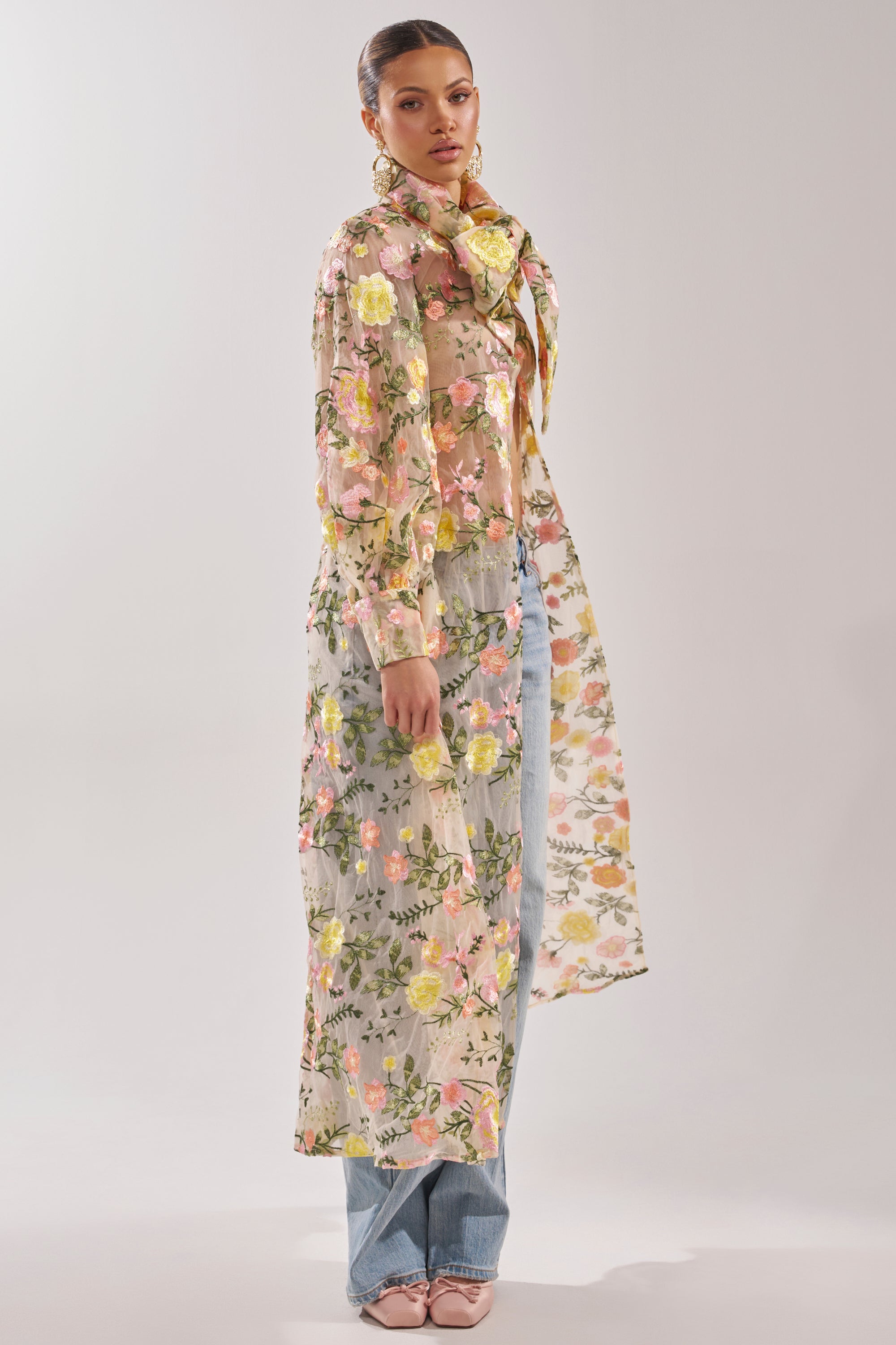 A woman wears the BOUQUET DREAMS FLORAL TULLE DUSTER with pastel floral embroidery, a matching scarf, light blue jeans, and pink shoes against a plain light background.