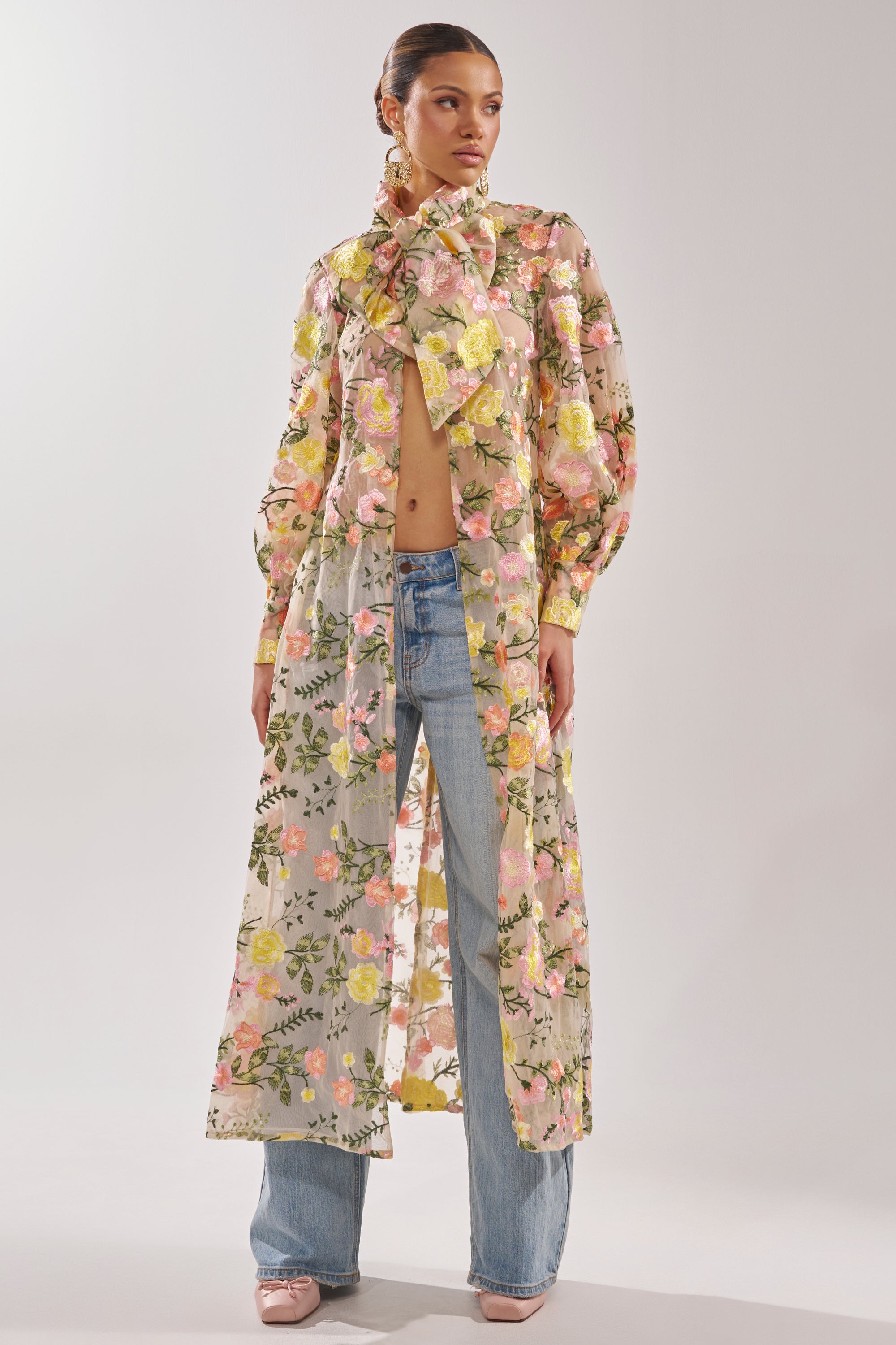 A woman poses in the BOUQUET DREAMS FLORAL TULLE DUSTER, a sheer maxi piece with colorful floral and leaf designs and a large neck bow, styled with blue jeans, pink shoes, and her hair in a sleek bun.