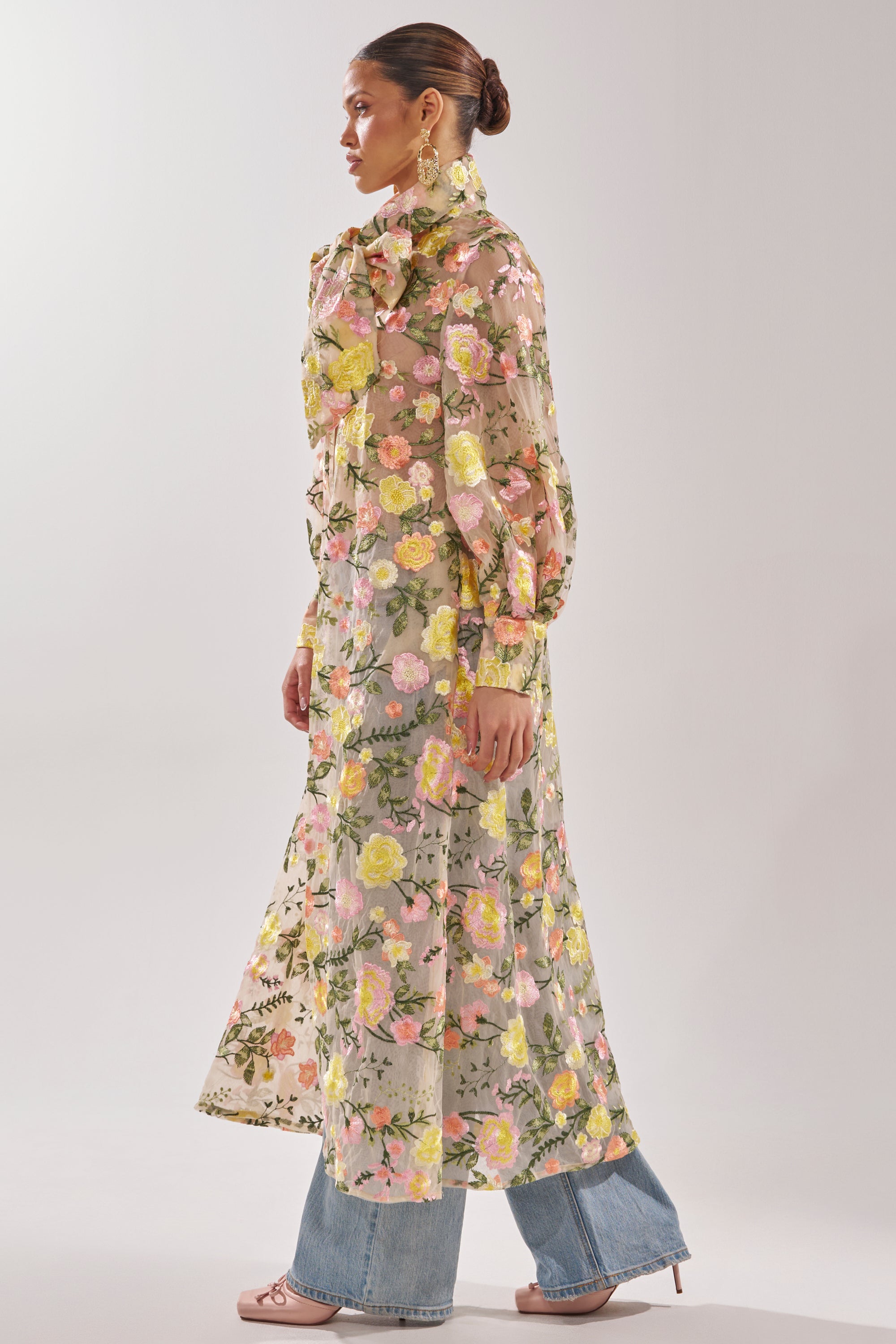 A woman stands sideways against a plain background, wearing the BOUQUET DREAMS FLORAL TULLE DUSTER with pink and yellow flowers over blue jeans, paired with hoop earrings, a bun hairstyle, and light pink heels.