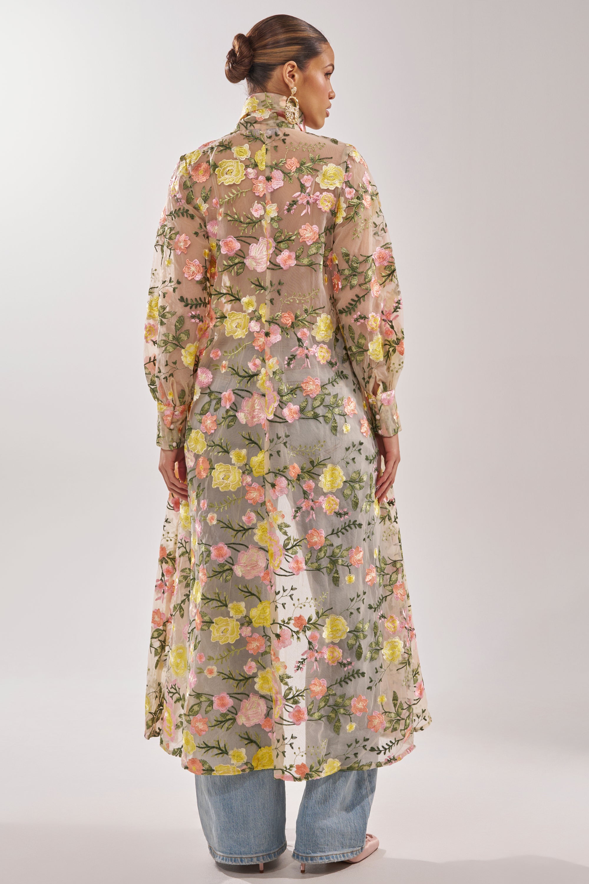 A woman with a sleek bun faces away, wearing the BOUQUET DREAMS FLORAL TULLE DUSTER—sheer with pink, yellow, and green florals—layered over blue jeans and heels. The background is plain and light.