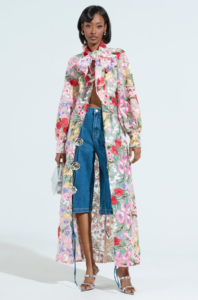 A person models the GARDEN PARTY FLORAL APPLIQUE LONG BLOUSE, featuring a bow neckline and worn over cropped jeans with decorative buttons, paired with a silver handbag and metallic sandals. The ensemble is set against a plain white background.