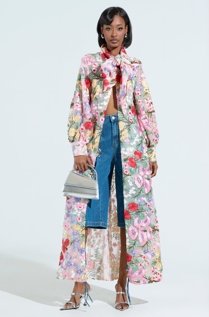 A person in a vibrant Garden Party Floral Applique Long Blouse over denim shorts pairs it with a sheer mesh top, carries a small silver handbag, and wears strappy silver heels against a plain white backdrop.