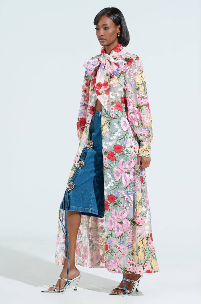 A woman models the Garden Party Floral Applique Long Blouse, featuring a large bow, paired with a longline coat and buttoned denim midi skirt. Her outfit is finished with strappy high heels, set against a plain backdrop.