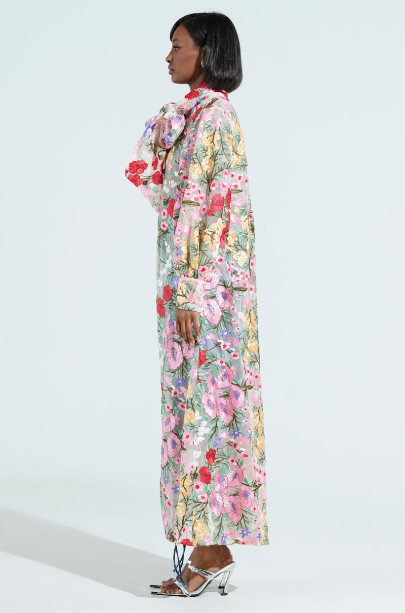 A woman poses in profile against a light backdrop, wearing the GARDEN PARTY FLORAL APPLIQUE LONG BLOUSE with vibrant pink, red, and green floral prints. She has a short bob hairstyle, open-toed silver heels, sheer mesh panels in her attire, and a large bow around her neck.