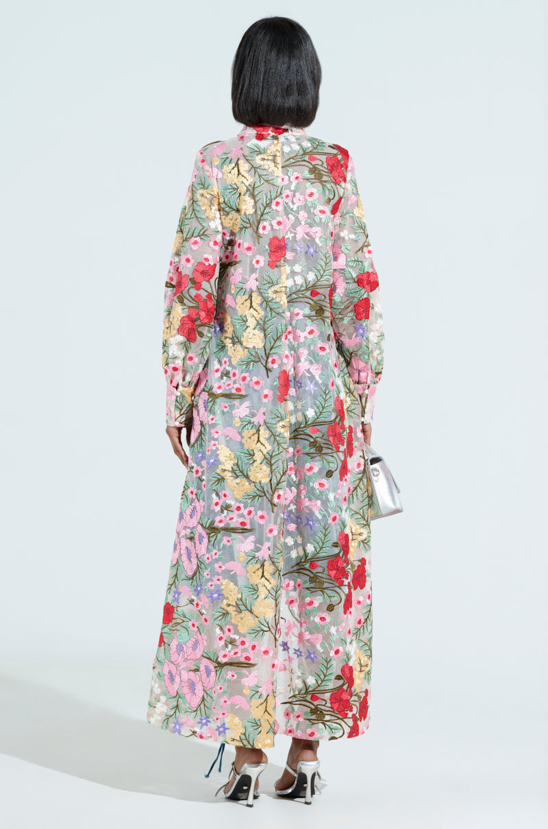 A person with shoulder-length hair stands facing away, wearing the GARDEN PARTY floral applique long blouse, exuding charm and grace. They complement the look with a small silver handbag and high-heeled shoes for added sophistication.