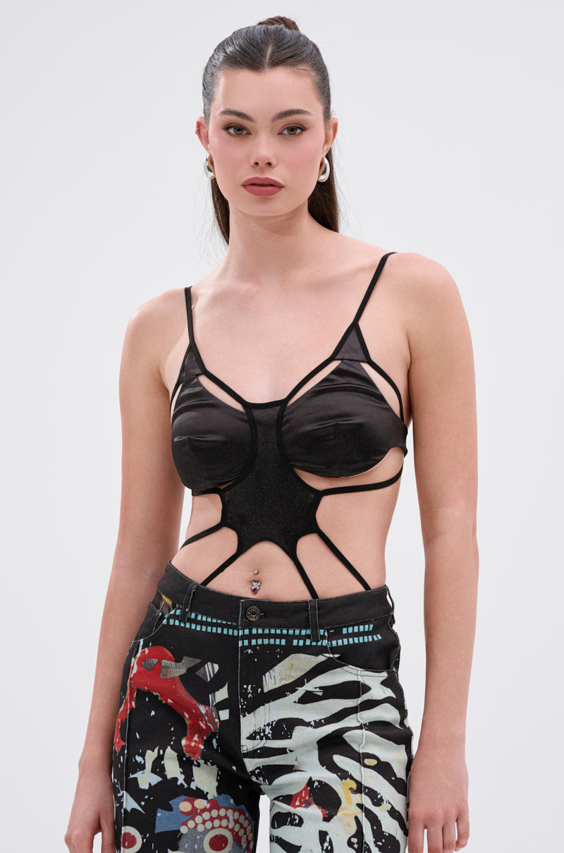 A woman with long dark hair in a ponytail wears the RISKY MOVES CUTOUT SLEEVELESS BODYSUIT and bold patterned pants, standing against a plain light background and looking confidently at the camera.