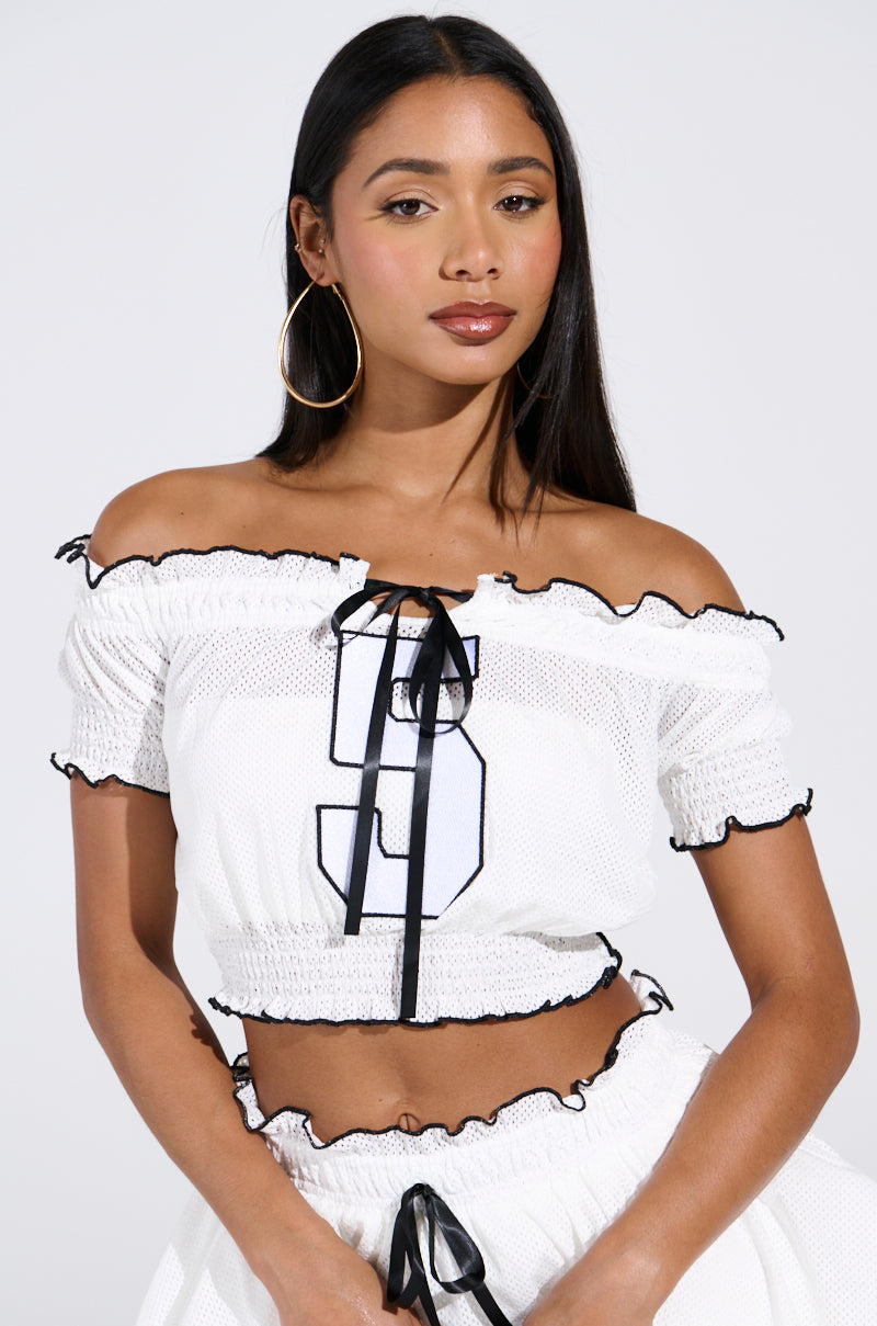 A woman with long straight hair and large hoop earrings wears the white LINDSEY CROPPED TOP, featuring ruffled edges, black ribbon accents, mesh jersey fabric, a bold number 9 on the front, paired with a matching white skirt.
