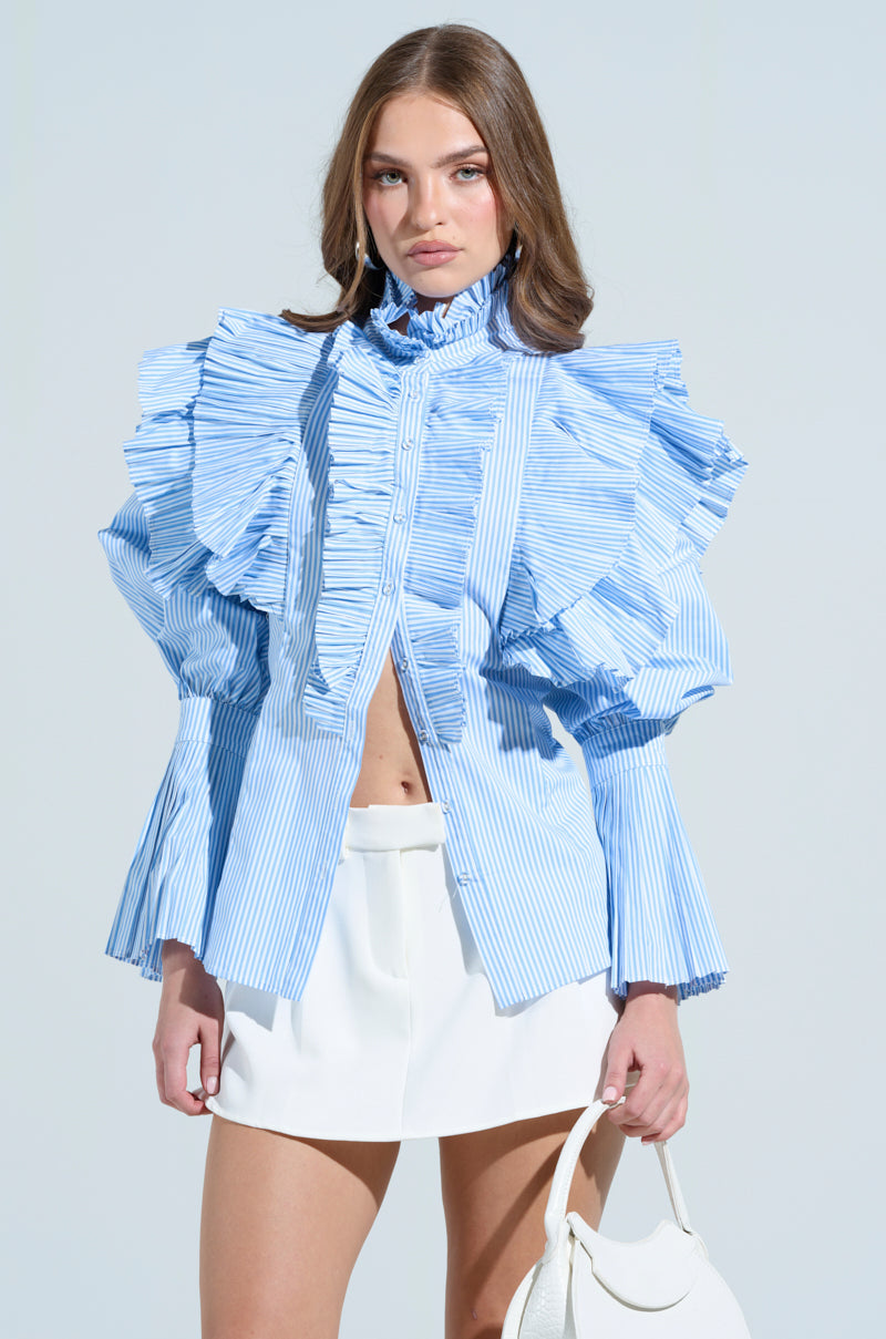 Someone with long hair dons the KAREN BUTTON DOWN BLOUSE, characterized by blue and white stripes and pleated ruffles on large sleeves and collar, paired with a white skirt. They carry a white bag against a plain backdrop.