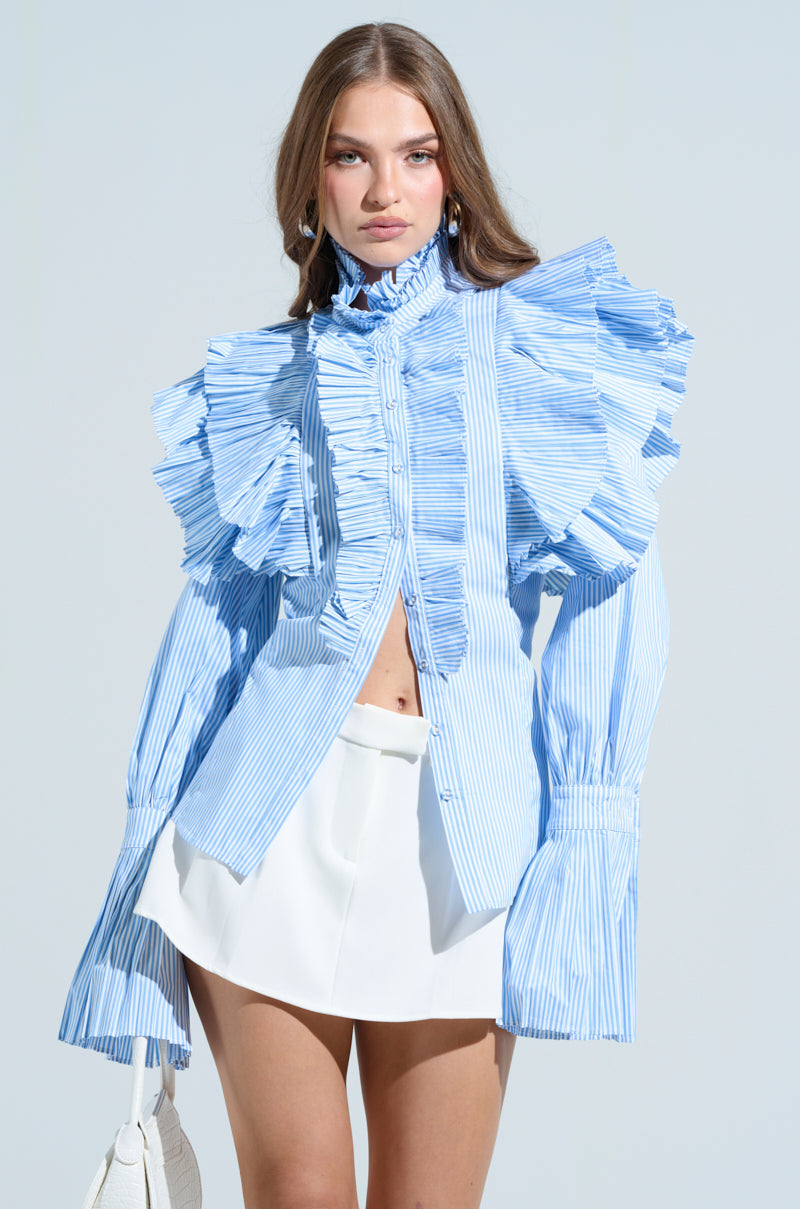 A woman wears the KAREN BUTTON DOWN BLOUSE, blue and white with large pleated sleeve ruffles, paired with a white skirt. She holds a small white bag and stands against a light background, gazing directly at the camera.
