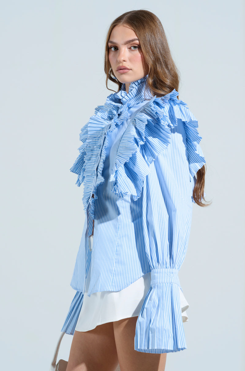 Wearing the KAREN BUTTON DOWN BLOUSE, featuring pleated ruffles and a blue and white striped pattern, a person with long hair pairs it with a white skirt. They stand against a light blue background, gazing to the side.