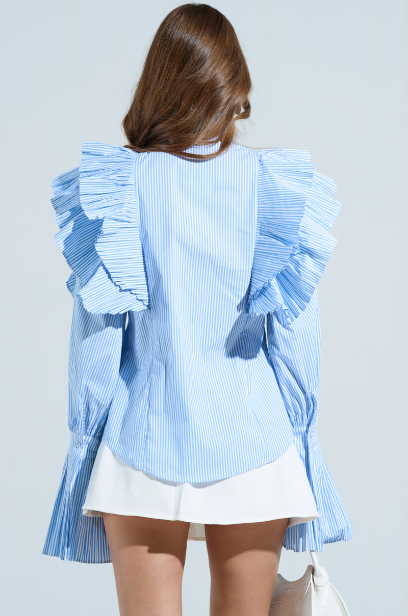 A person with long brown hair wears the KAREN BUTTON DOWN BLOUSE, blue and white with pleated ruffles, paired with white shorts. They hold a white bag against a light blue background.