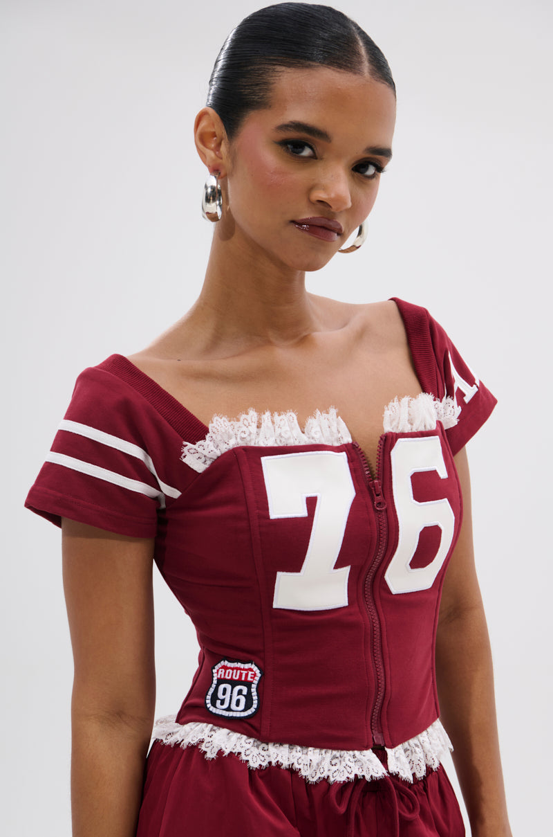 A woman wears the PLAY TIME IS OVER FRONT ZIP CORSET TOP in maroon with white lace trim, large "76" on the front, striped sleeves, and a "Route 96" patch, styled with hoop earrings against a plain light background. 1