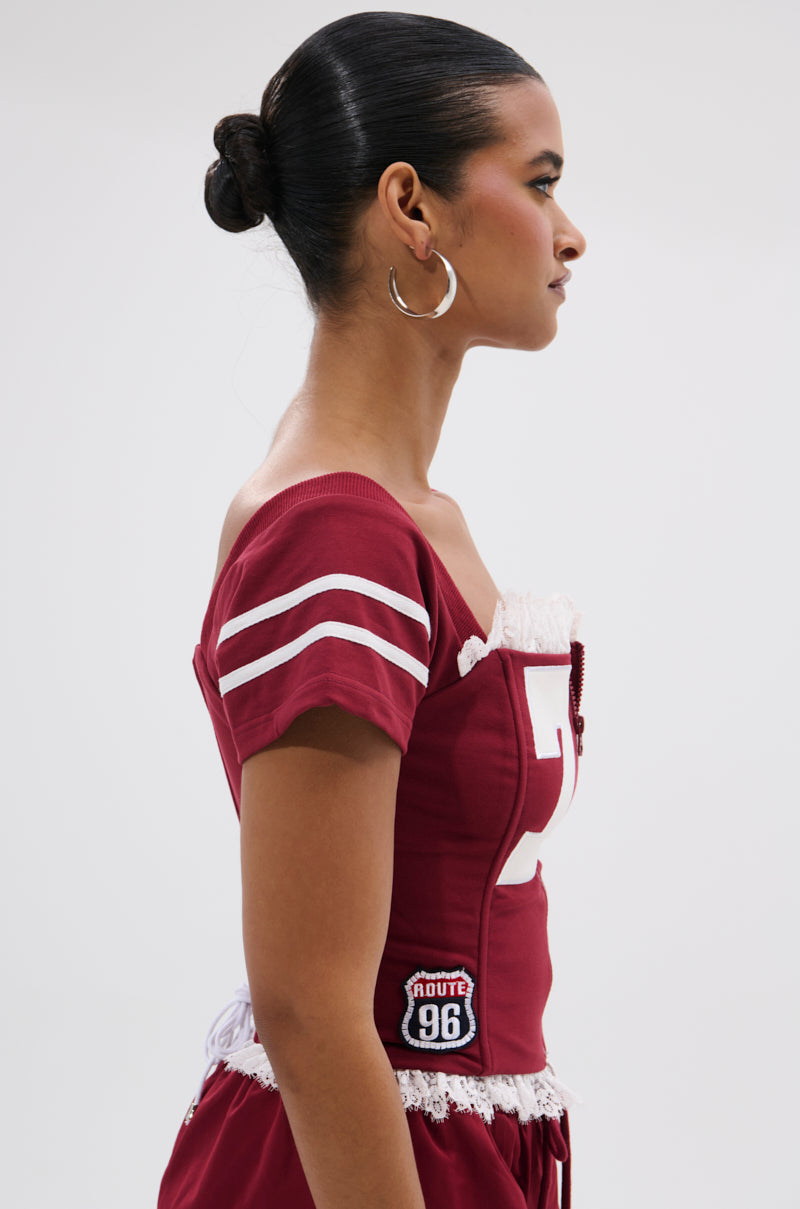 A woman with dark hair in a bun wears the PLAY TIME IS OVER FRONT ZIP CORSET TOP—an off-shoulder maroon sporty style with white sleeve stripes and a "Route 96" patch—paired with large hoop earrings, shown in profile against a plain background. 2