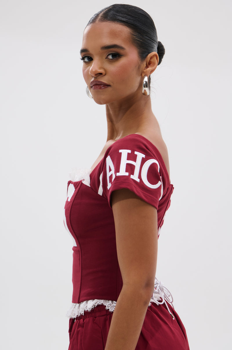 A woman with a sleek bun wears the PLAY TIME IS OVER FRONT ZIP CORSET TOP in maroon with white lace trim and bold white sleeve lettering, confidently looking over her shoulder against a plain background. 3