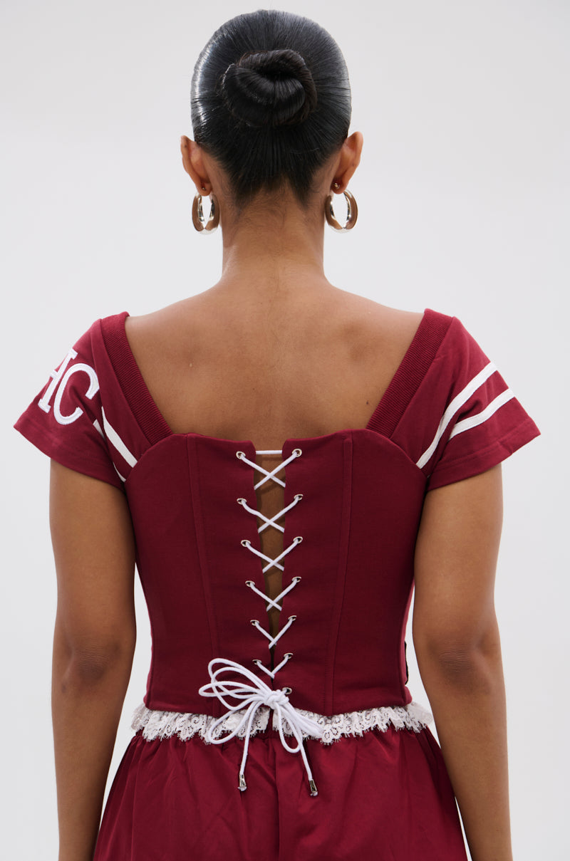A person with dark hair in a bun wears the PLAY TIME IS OVER FRONT ZIP CORSET TOP—a maroon, cropped corset-style top with crisscross lacing, short sleeves, white trim, and lace at the waist for an elegant look. 4