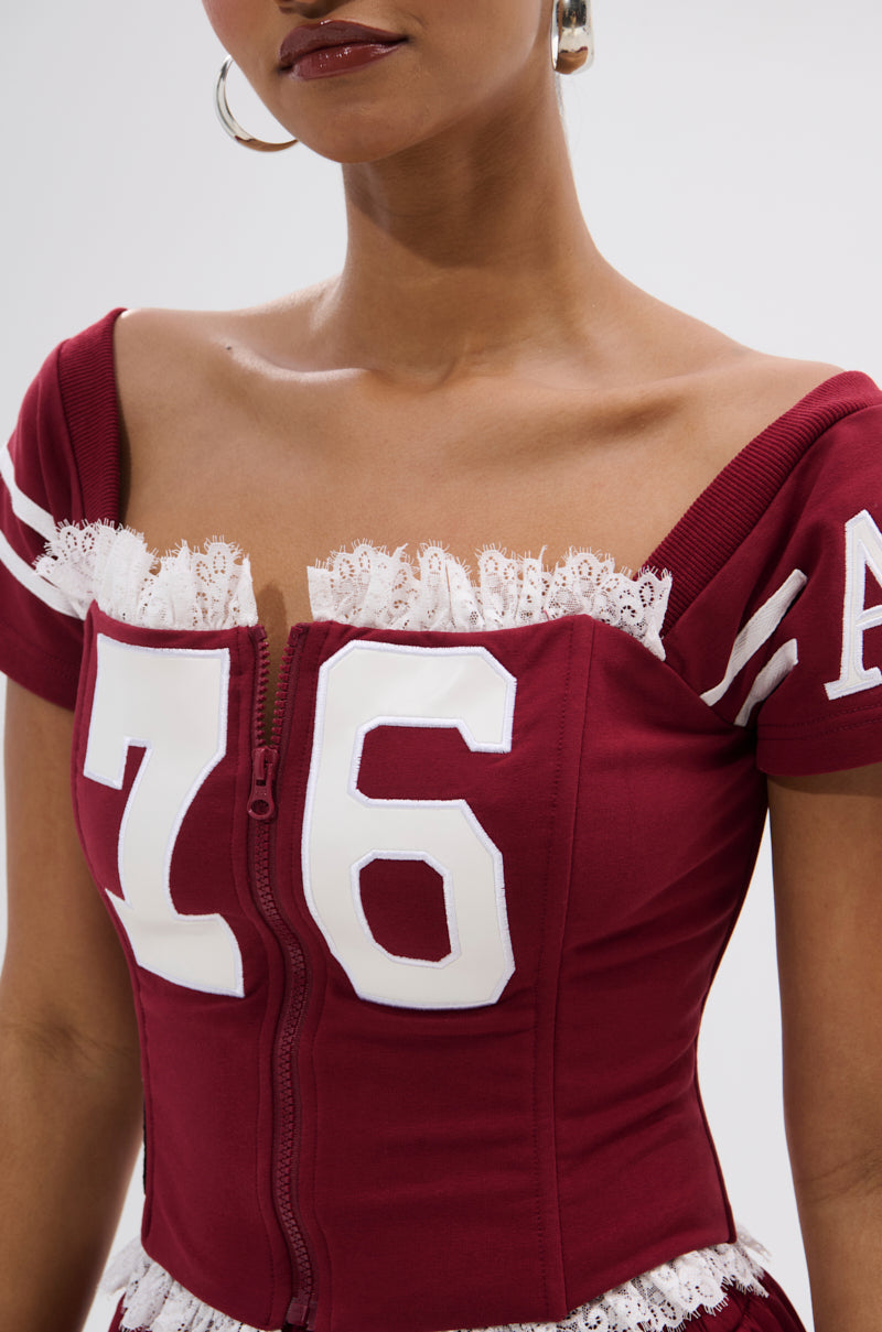 A woman wears the PLAY TIME IS OVER FRONT ZIP CORSET TOP in maroon, featuring sporty white lace trim, sleeve stripes, and a bold '76' on the front. Paired with hoop earrings and red lipstick, she achieves a chic athletic-inspired style. 7