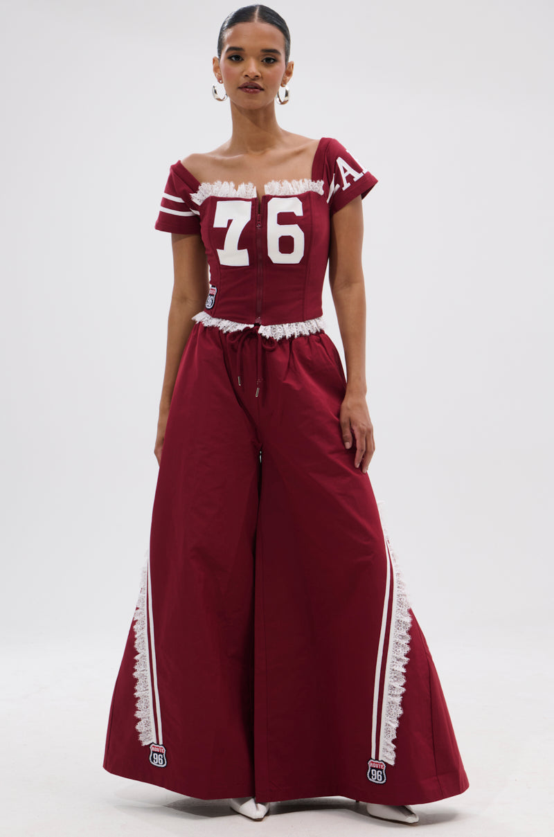 A woman poses confidently in PLAY TIME IS OVER OVERSIZED TRACK PANT, a sporty wide-leg style with white lace trim and athletic accents, paired with a maroon off-shoulder top featuring the number 76 against a plain white background. 0
