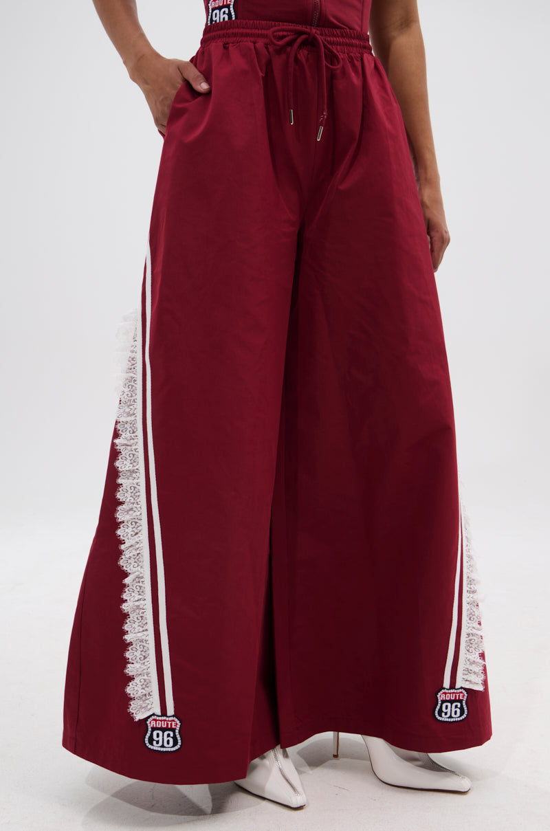 Wearing the PLAY TIME IS OVER OVERSIZED TRACK PANT—wide-leg, high-waisted maroon pants with white lace trim, side stripes, and “Route 96” embroidered near the hem—styled with white heeled boots for a sporty look. 1
