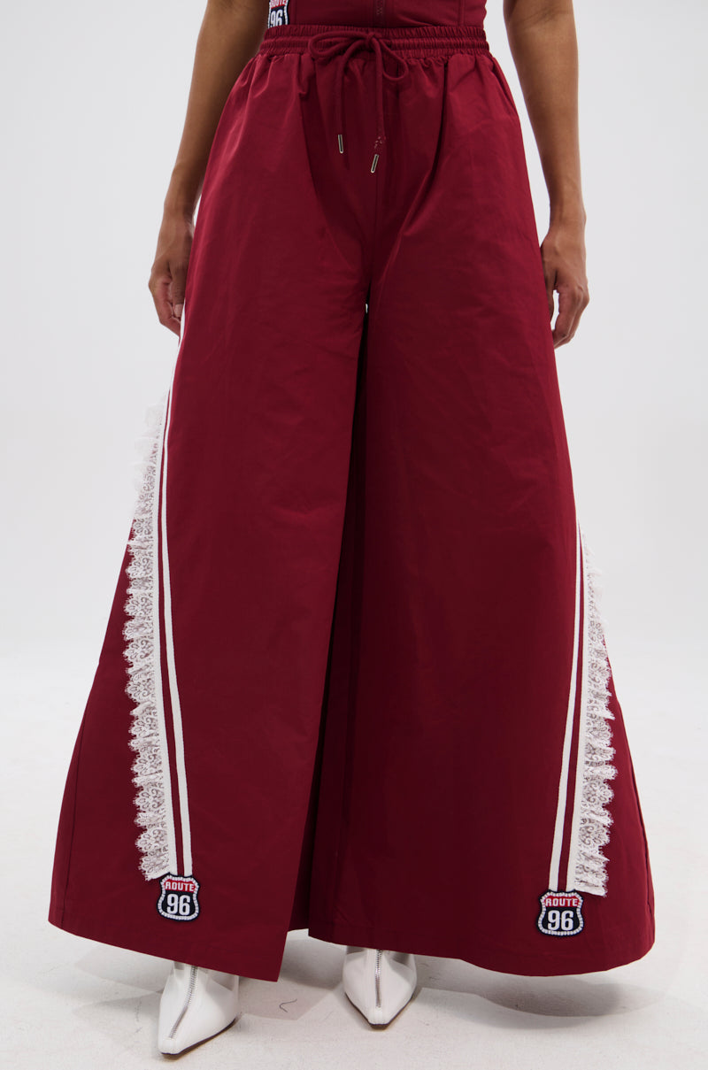 A person wears PLAY TIME IS OVER OVERSIZED TRACK PANT: maroon, wide-leg silhouette, white lace trim and vertical stripes along the sides, "1996" patches by the hem, styled with white pointed-toe shoes. 2