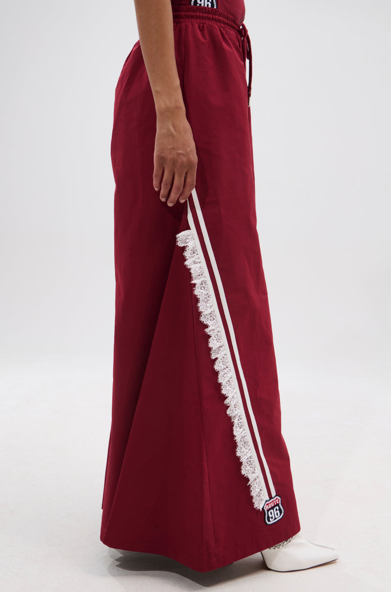 A person wears the PLAY TIME IS OVER OVERSIZED TRACK PANT, a long, deep red wide-leg track pant with white side stripes, lace trim, and a "96" patch near the hem, styled with white pointed-toe boots. 3