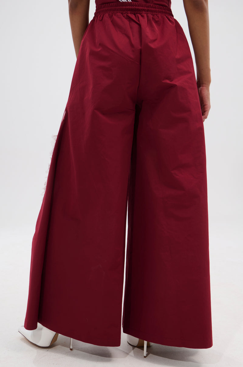 A person wearing the PLAY TIME IS OVER OVERSIZED TRACK PANT in maroon with white pointed-toe heels, shown from the back against a plain light background. 4