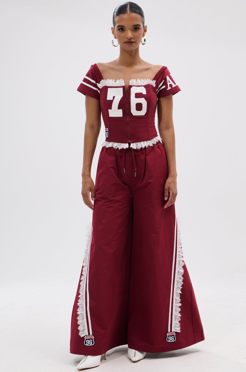 A woman stands facing forward in the PLAY TIME IS OVER OVERSIZED TRACK PANT, a maroon wide-leg silhouette with white lace trim and athletic patches, paired with a matching off-shoulder top numbered 76. Her hair is tied back and she wears white pointed shoes. 5