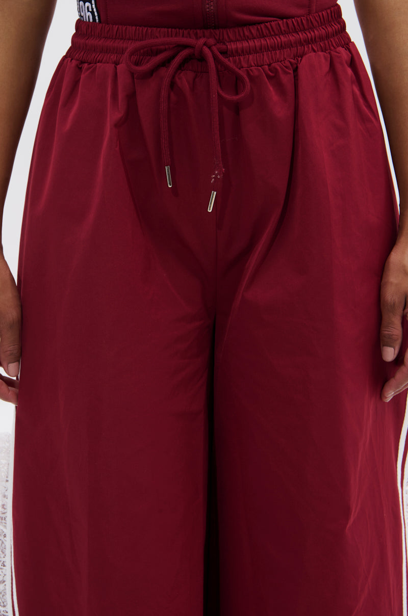 A person wearing the PLAY TIME IS OVER OVERSIZED TRACK PANT—high-waisted, loose maroon track pants with a drawstring waist, white side stripes, and wide-leg silhouette. Only their lower torso and hands are visible. 6