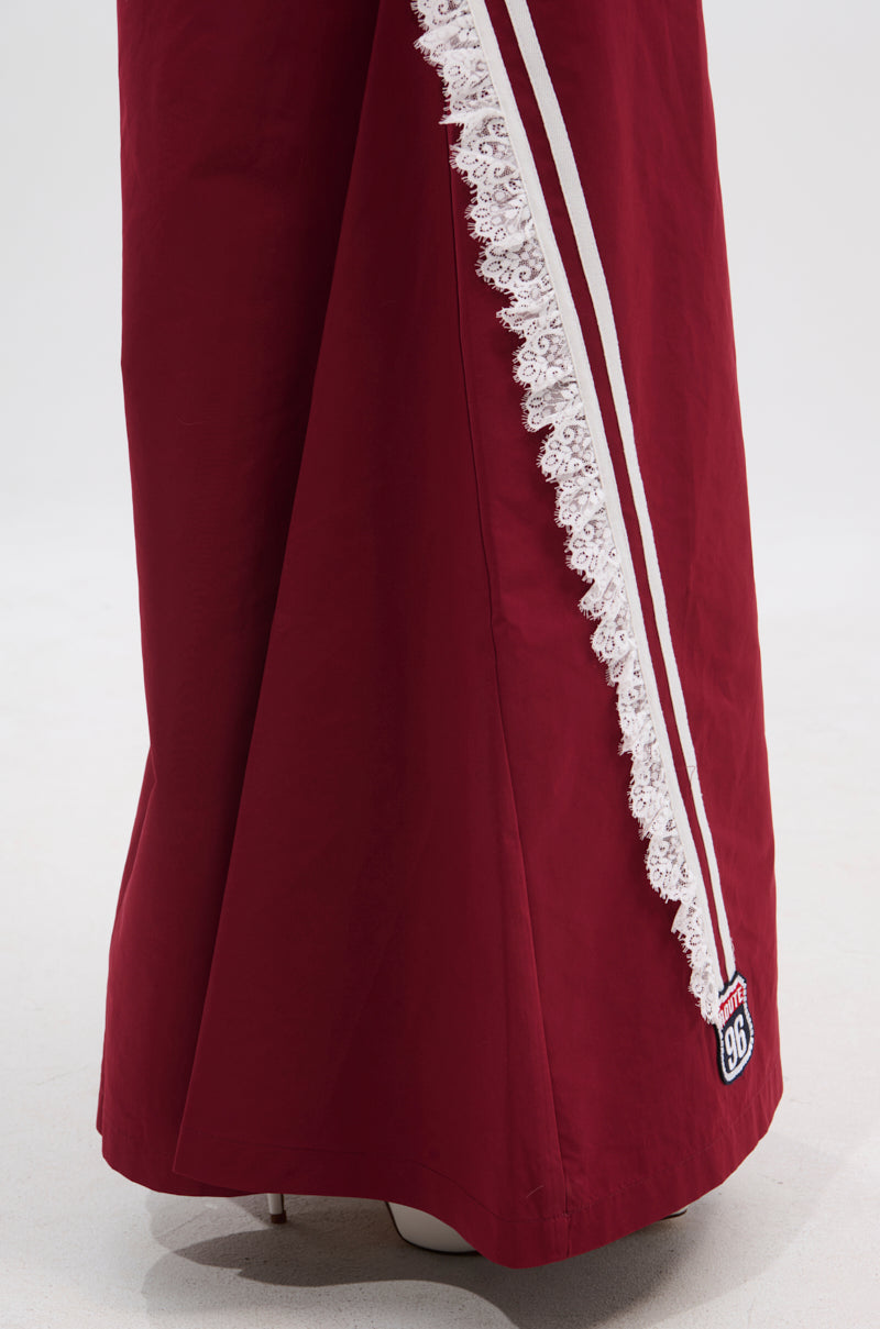 A close-up of the PLAY TIME IS OVER OVERSIZED TRACK PANT in maroon, featuring a white lace trim and diagonal stripe, sporty style, and a small "916" embroidered patch near the hem. The background is plain and light. 7