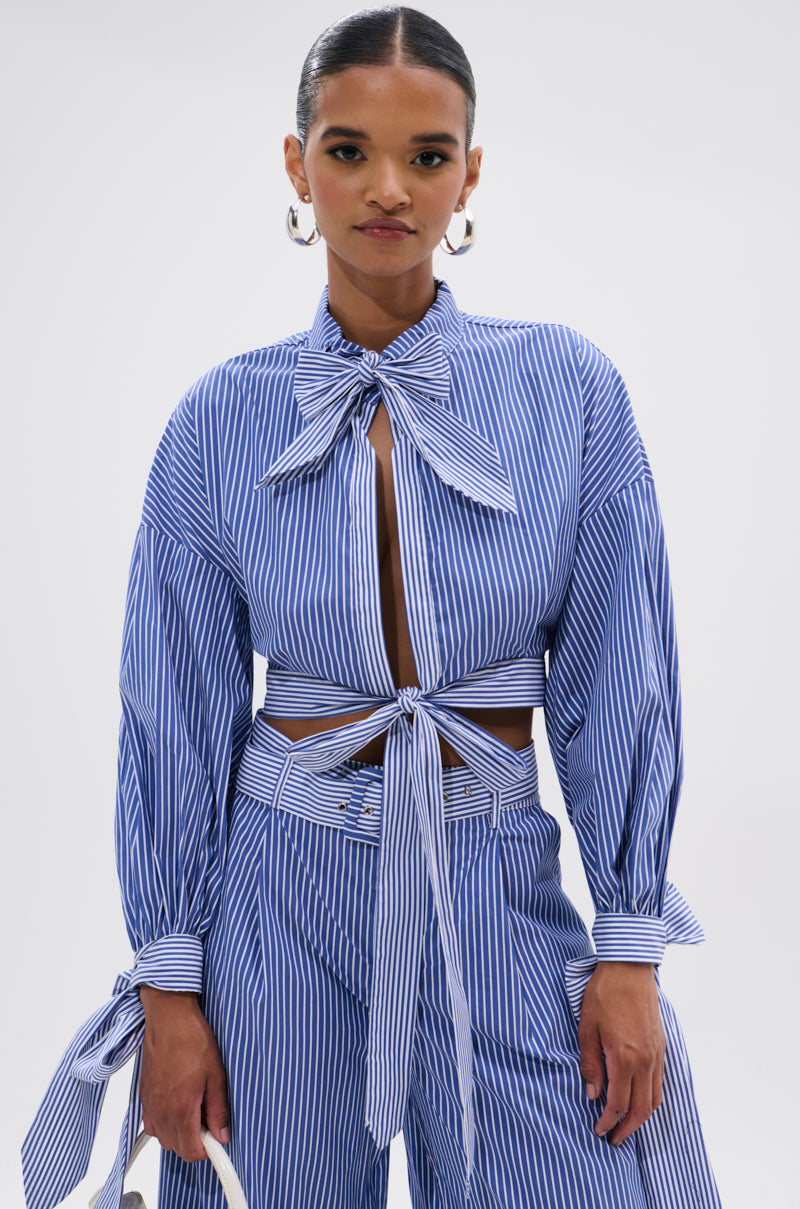 A woman models the UNDER THE SUN STRIPE TIE FRONT BLOUSE with blue-and-white stripes, a bow tie at the neck, and high-waisted pants. She wears hoop earrings, has slicked-back hair, and poses against a white background.