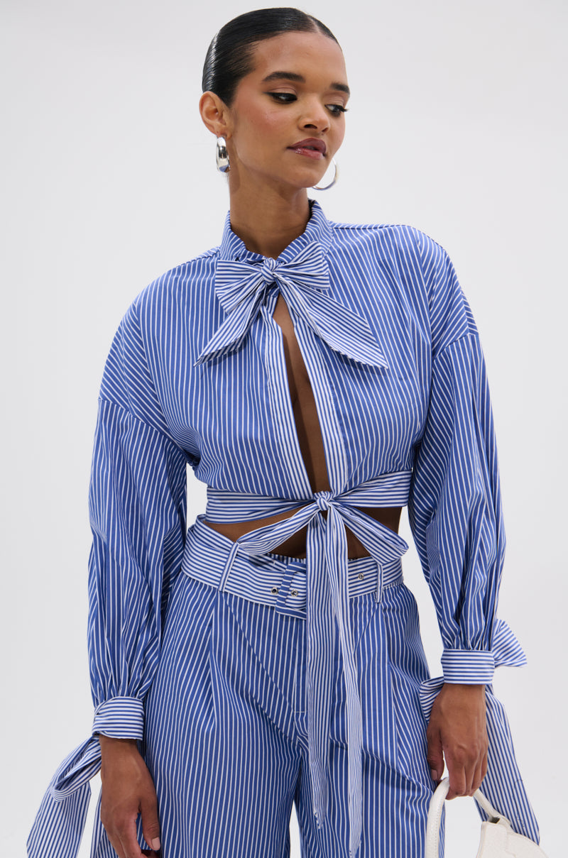A woman stands against a plain white background wearing the UNDER THE SUN STRIPE TIE FRONT BLOUSE, a blue-and-white striped top with a large collar bow, waist tie, and long sleeves, paired with high-waisted cropped pants.