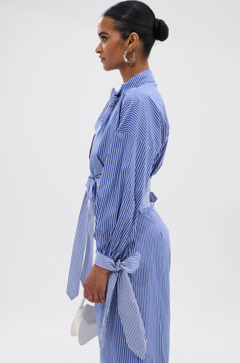 A woman stands in profile against a plain background, wearing the UNDER THE SUN STRIPE TIE FRONT BLOUSE with a waist tie and bow sleeve details, holding a light clutch, accessorized with hoop earrings and a sleek bun.
