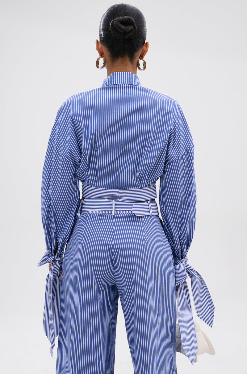 A person with their hair in a bun is seen from behind, wearing the UNDER THE SUN STRIPE TIE FRONT BLOUSE, featuring blue-and-white stripes and a cropped boxy silhouette that stands out against a plain white background.