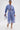 She wears the UNDER THE SUN STRIPE TIE FRONT BLOUSE, a blue-and-white striped, cropped, boxy silhouette top with matching wide-leg culotte shorts, accessorized with a small white purse and white pointed-toe heels.