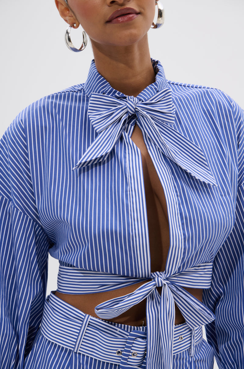 A woman wears the UNDER THE SUN STRIPE TIE FRONT BLOUSE—blue-and-white stripes, oversized bows, long sleeves, and a cut-out front—with matching pants and large hoop earrings for a stylish cropped boxy look.
