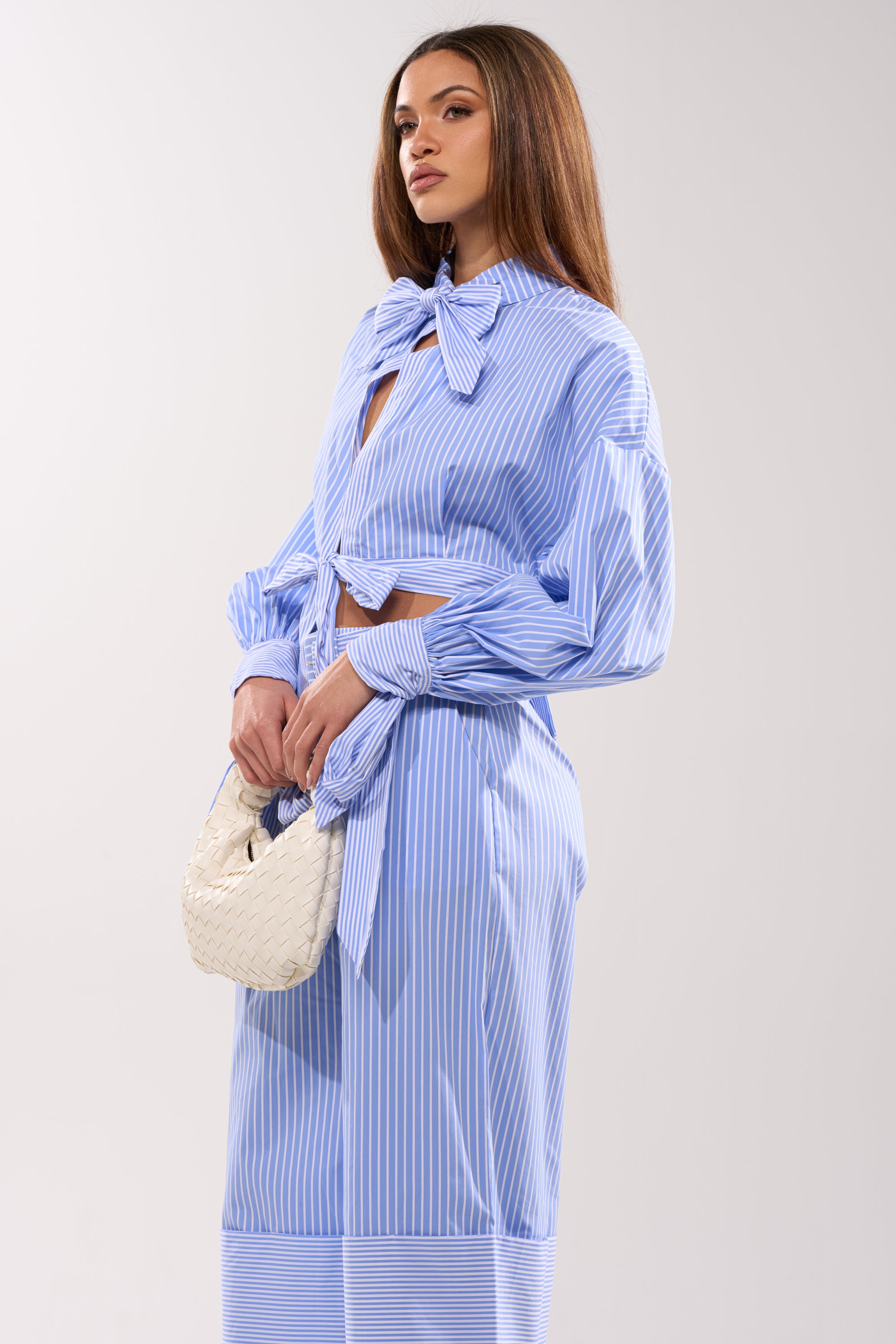 A woman stands against a light background wearing the NOT LIKE ANYONE ELSE POPLIN BLOUSE, featuring blue and white stripes with a large neck bow, paired with a matching skirt and holding a white woven handbag.