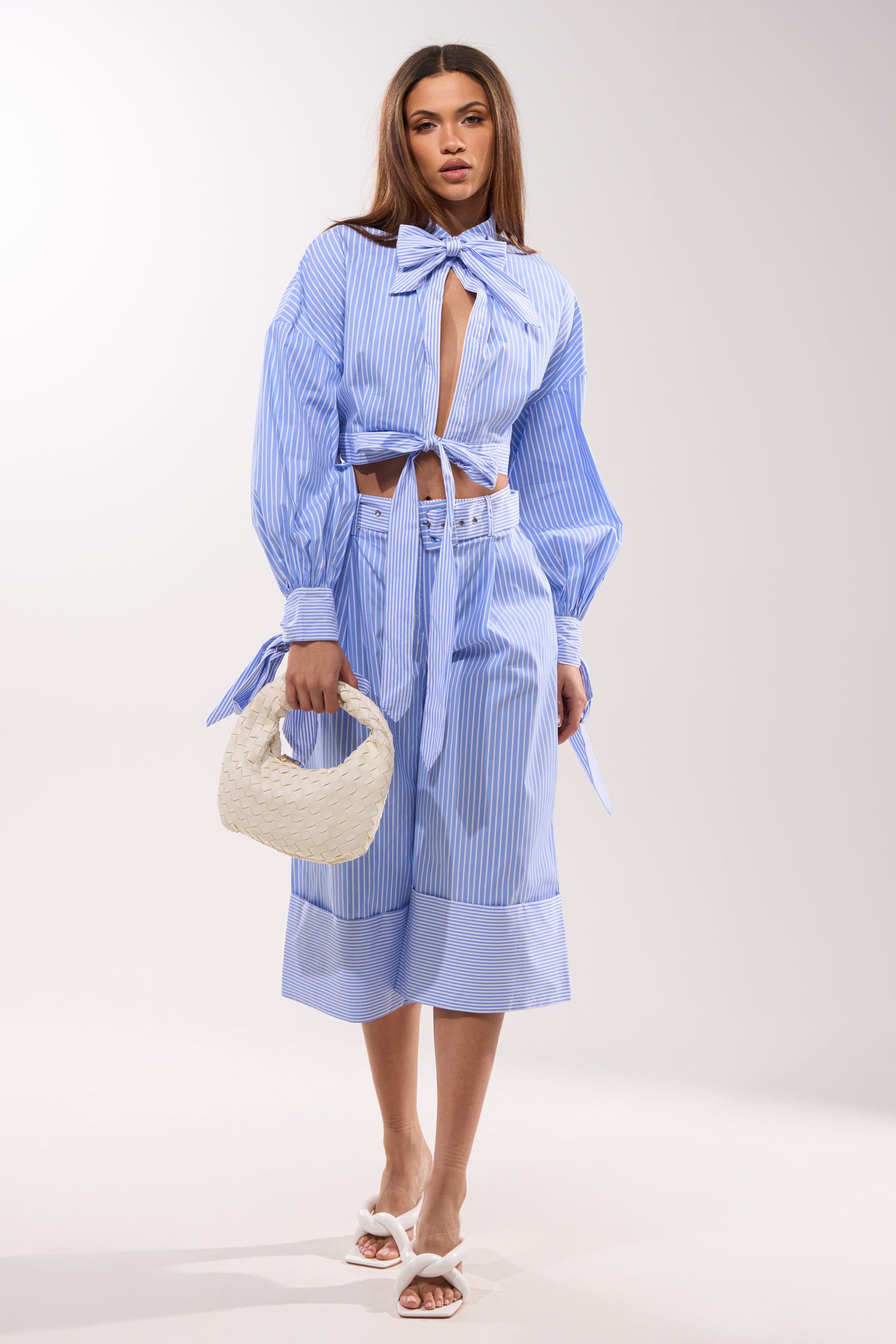 A woman poses in the NOT LIKE ANYONE ELSE POPLIN BLOUSE—blue and white striped with a large bow and front cutout—paired with a matching belted midi skirt, white woven handbag, and white heeled sandals against a plain background.