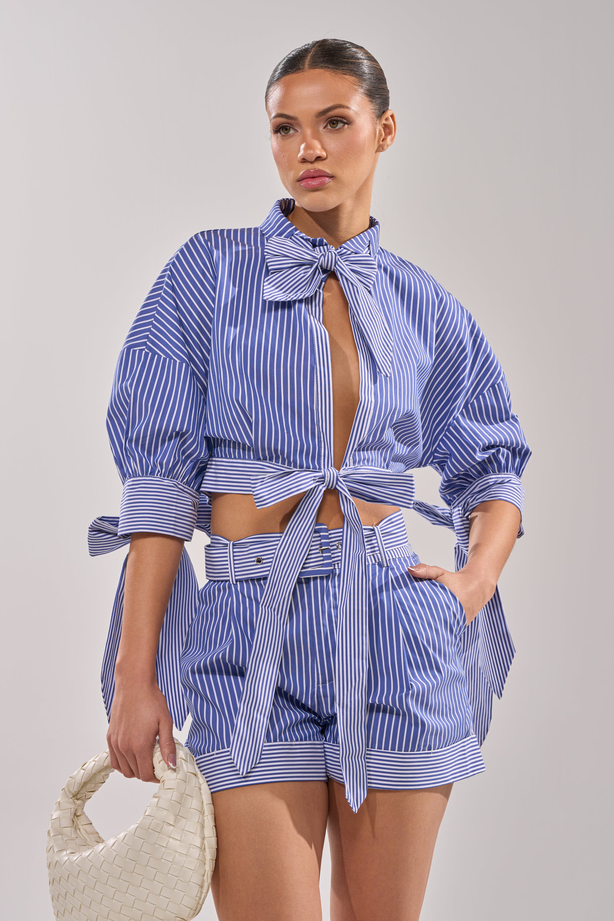 A woman wears the GUESS WHO POPLIN BLOUSE, a blue and white pinstripe matching set with a tied cropped shirt, high-waisted shorts, and a large bow neckline, paired with a white woven handbag.
