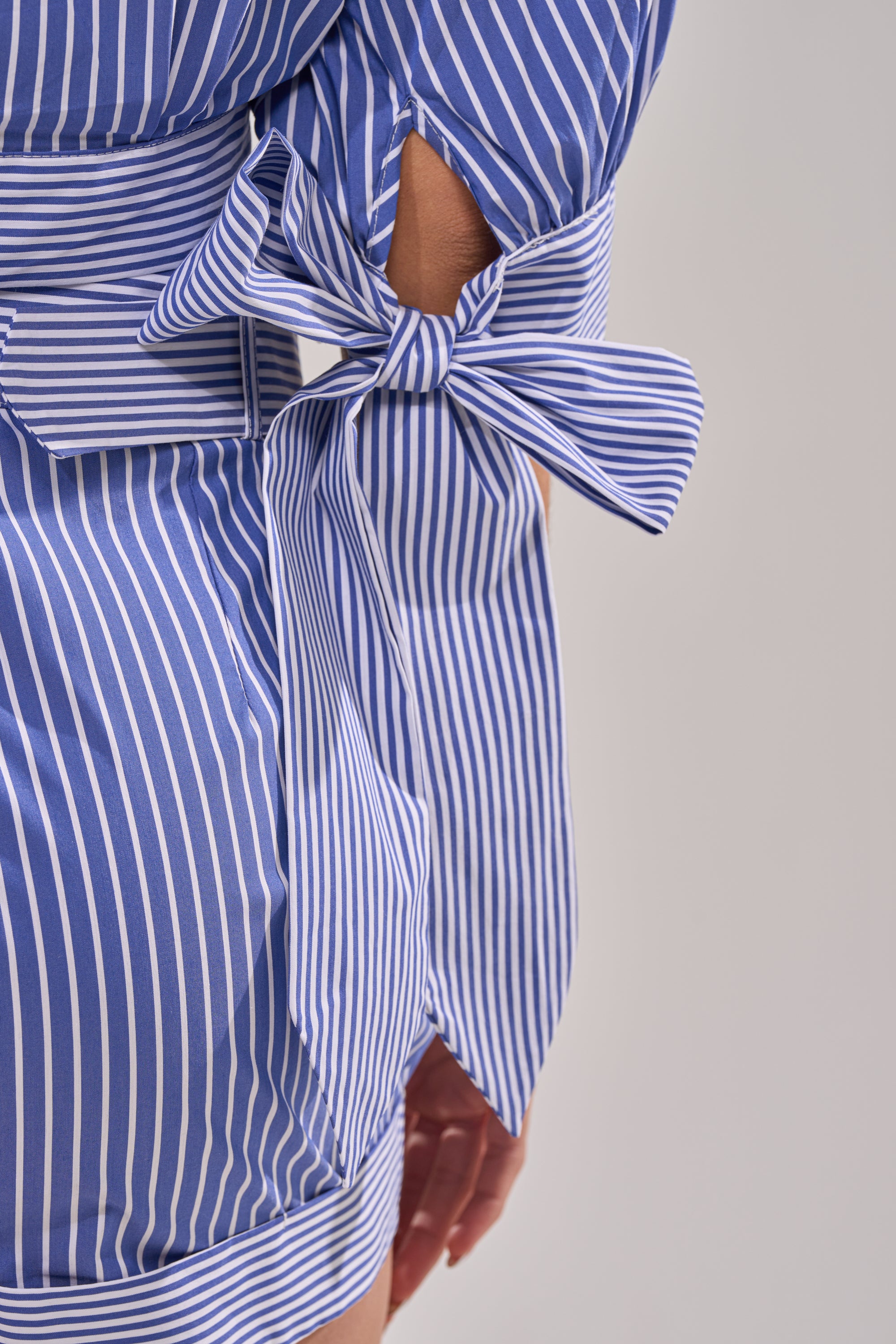 A close-up of a person wearing the GUESS WHO POPLIN BLOUSE featuring blue and white pinstripes, a large bow tied at the waist, and their hand resting at their side.