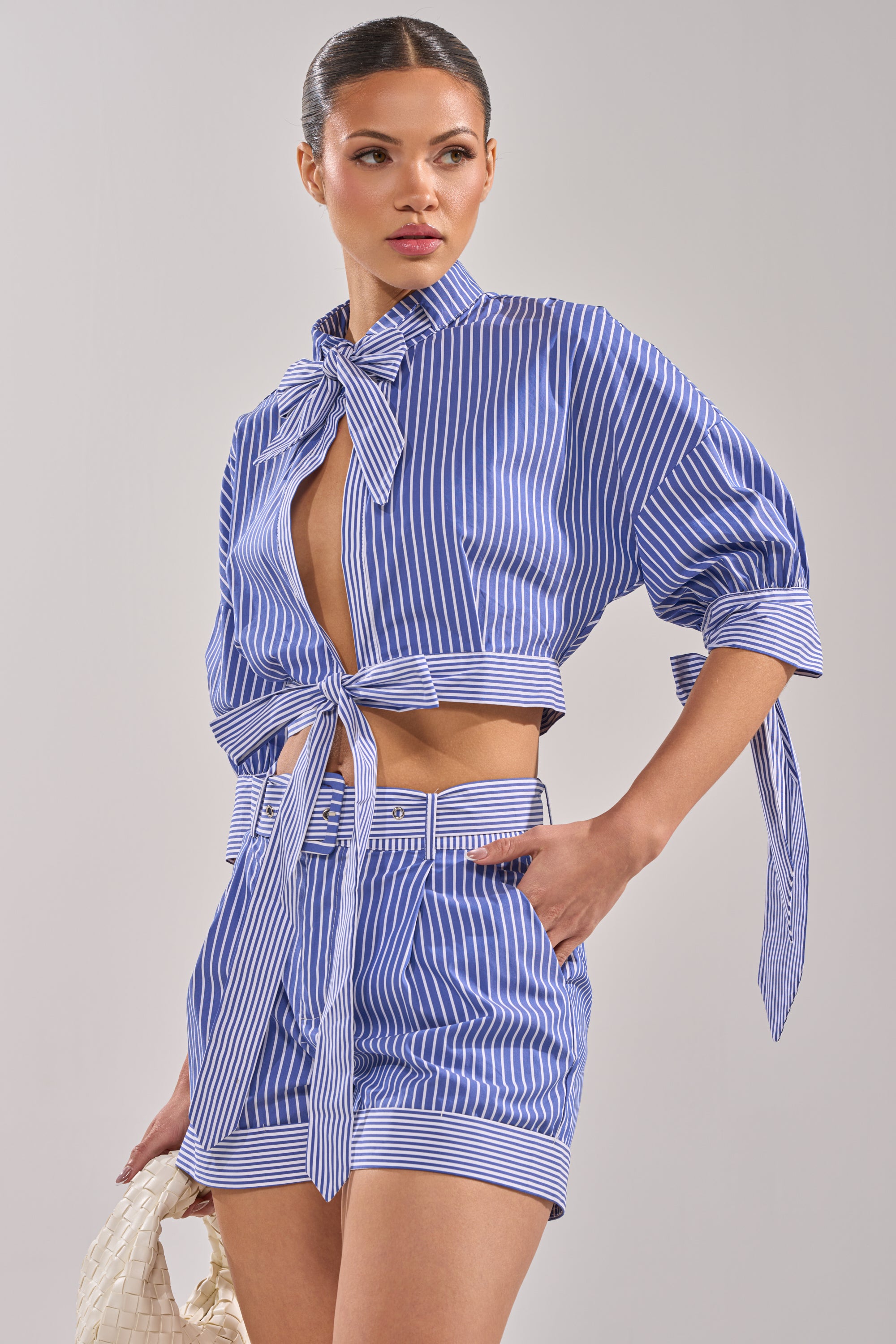 A woman models the GUESS WHO POPLIN BLOUSE—blue and white pinstripe, tie-front, cropped wrap style—with high-waisted shorts and a woven white bag, posing against a plain background.