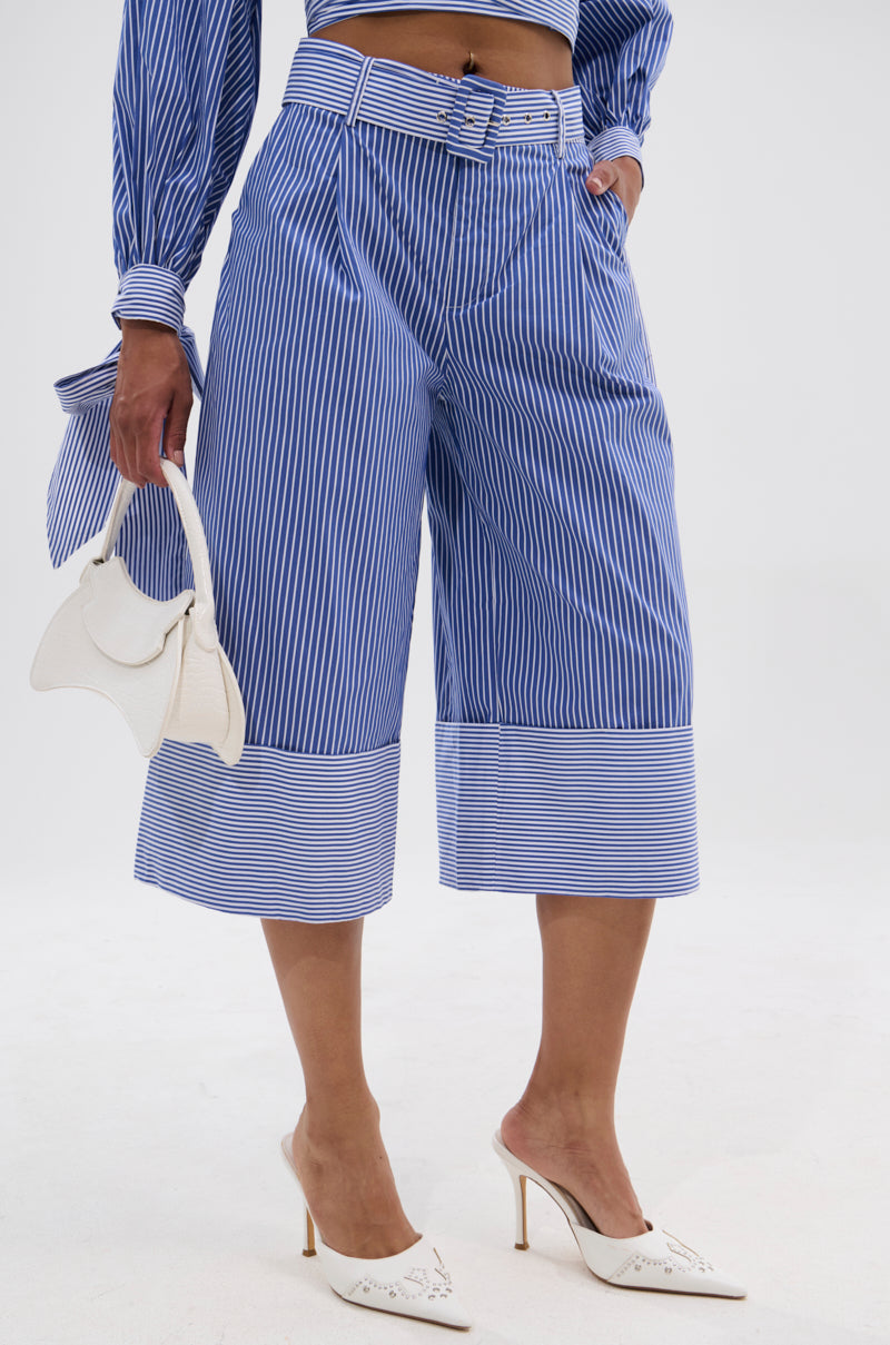 A person wearing UNDER THE SUN STRIPE SHORT, a matching striped long-sleeve top, white pointed-toe heels, and holding a small white handbag stands against a plain background.