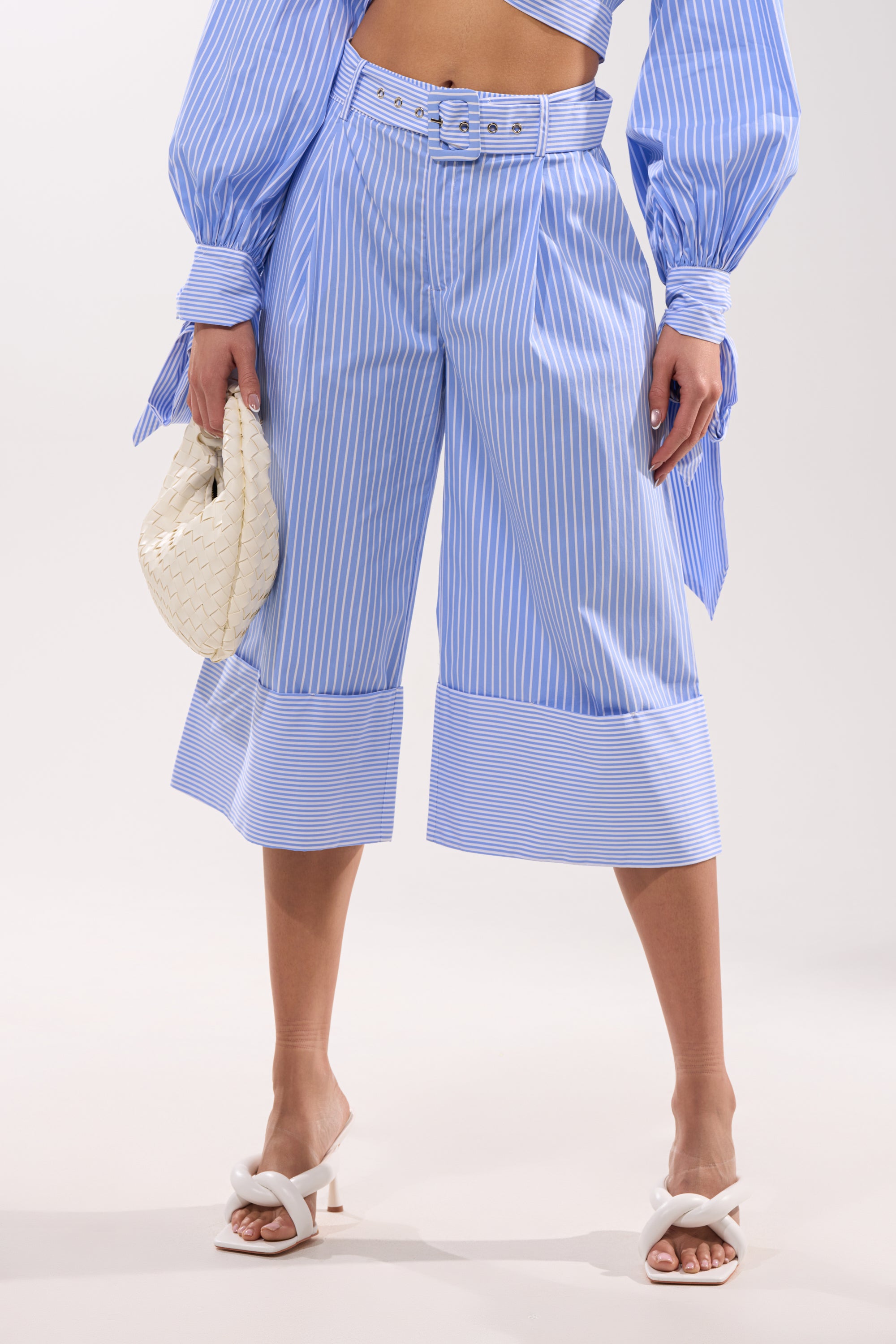 A person wears the NOT LIKE ANYONE ELSE POPLIN SHORT, a blue-and-white pinstripe high-rise short with a matching belted top, white heeled sandals, and holds a woven white handbag for an airy, stylish look.