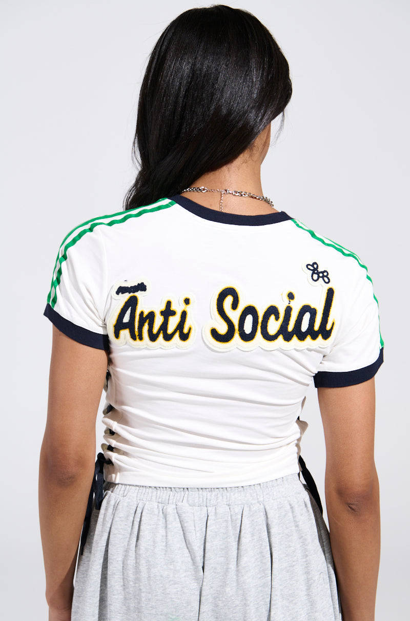 A person with long dark hair, seen from behind, wears the PATCH FOR DAYS BABY TEE—a cropped tee with green striped sleeves, bold “Anti Social” lettering on the back, lace-up sides—paired with a gray skirt.