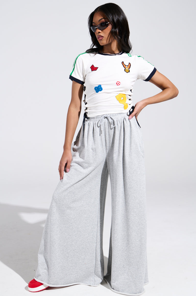 A woman stands confidently against a white background wearing gray wide-leg sweatpants, red shoes, sunglasses, and the PATCH FOR DAYS BABY TEE featuring colorful embroidered patches.