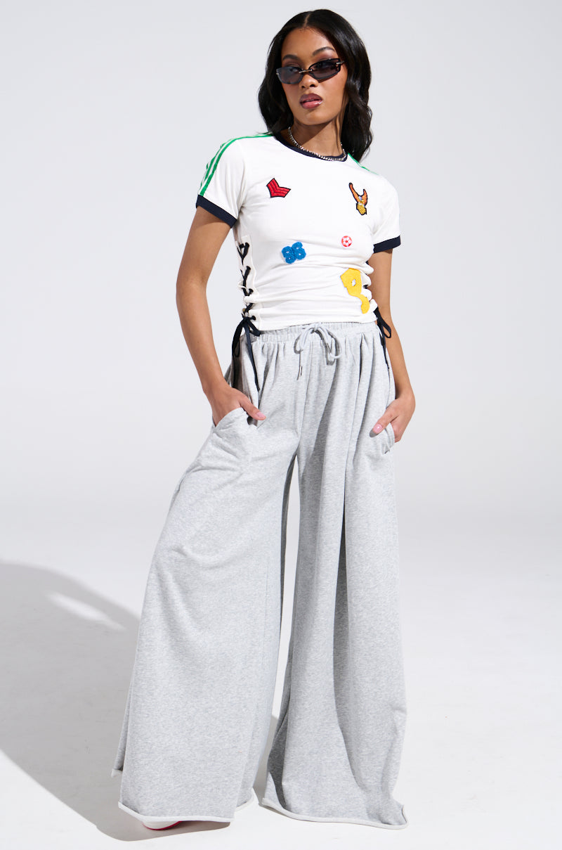 A woman stands confidently with her hands in her pockets, wearing sunglasses and the PATCH FOR DAYS BABY TEE paired with loose, light gray wide-leg pants against a plain background.