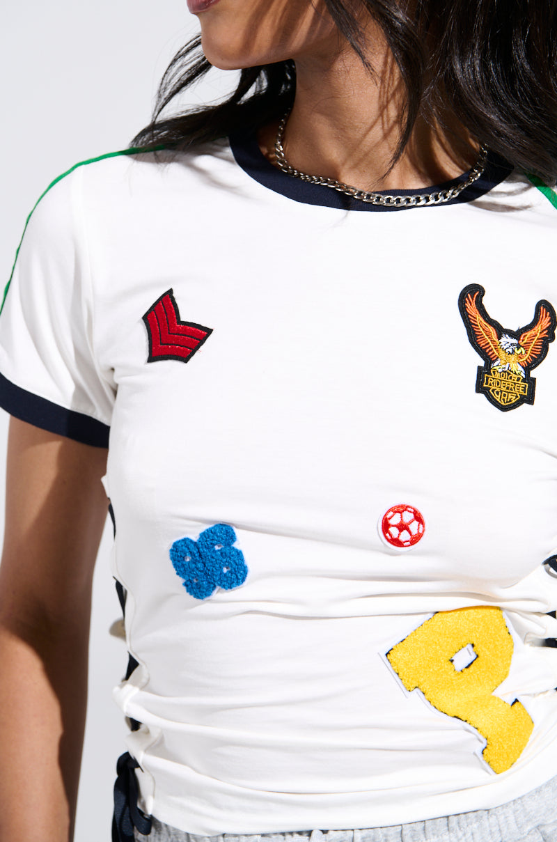 A woman wears the PATCH FOR DAYS BABY TEE, a cropped top with black trim, colorful embroidered patches like an eagle, 9, blue flower, and red chevron. She has wavy dark hair, a silver chain necklace, and trendy lace-up sides.