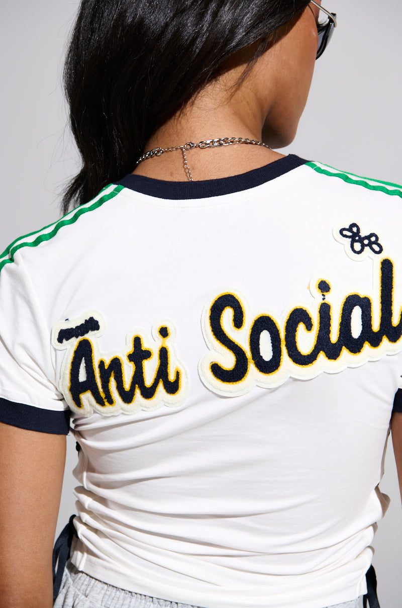 A person with long dark hair, facing away from the camera, wears the PATCH FOR DAYS BABY TEE—a cropped top featuring green shoulder stripes and “Anti Social” in bold yellow and black letters on the back.