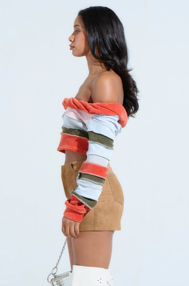 A person model showcases a MULTI KNIT CROPPED SWEATER featuring an off-the-shoulder, multicolored striped design reminiscent of patchwork. It's paired with a tan skirt, knee-high white boots, and a small handbag against a light blue background.
