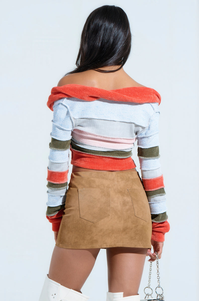 A woman faces away, wearing an off-the-shoulder MULTI KNIT CROPPED SWEATER and a tan suede skirt. Her look is finished with white knee-high boots and a small handbag. The plain light background accentuates the vibrant stripes of her outfit.