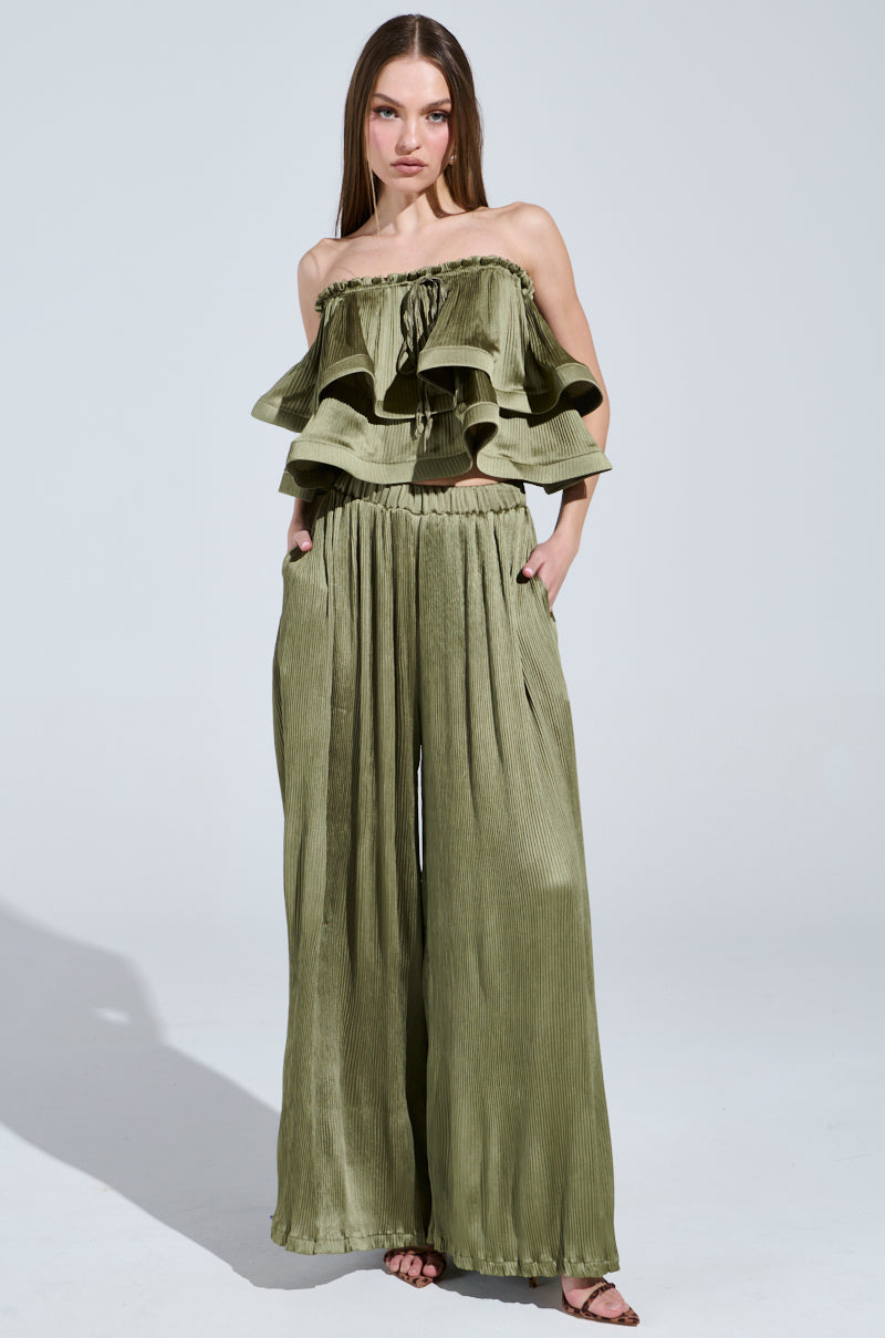 A person models a strapless khaki ruffle top with the "Fly Away With Me Pleated Palazzo Pant," featuring wide legs and pockets. They're accessorized with sandals, set against a plain light-colored background to accentuate the outfit's elegance.