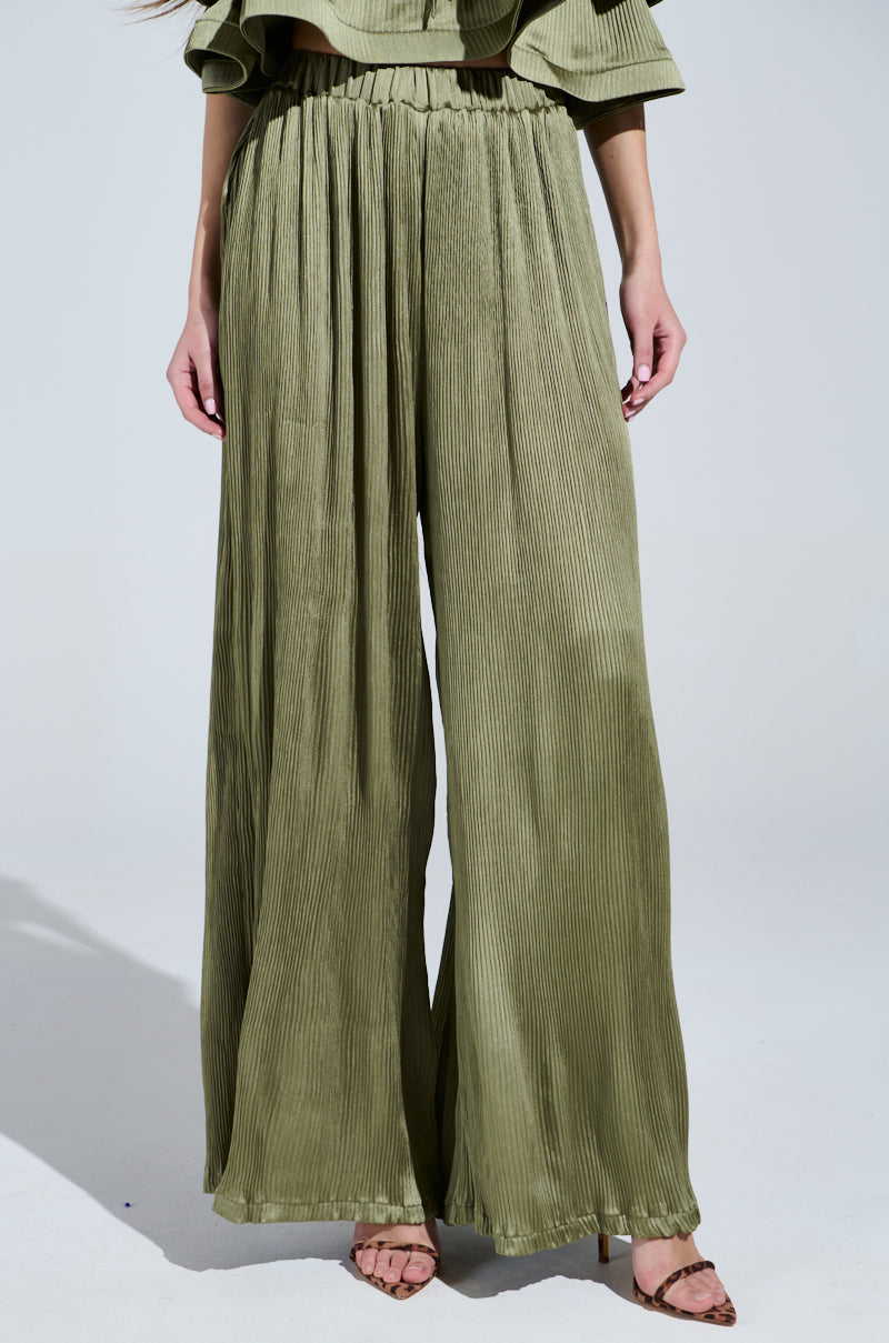 Wearing the FLY AWAY WITH ME PLEATED PALAZZO PANT in olive green, paired with a matching top and elegant brown open-toed shoes, the wide-leg silhouette is perfectly showcased against a plain background.